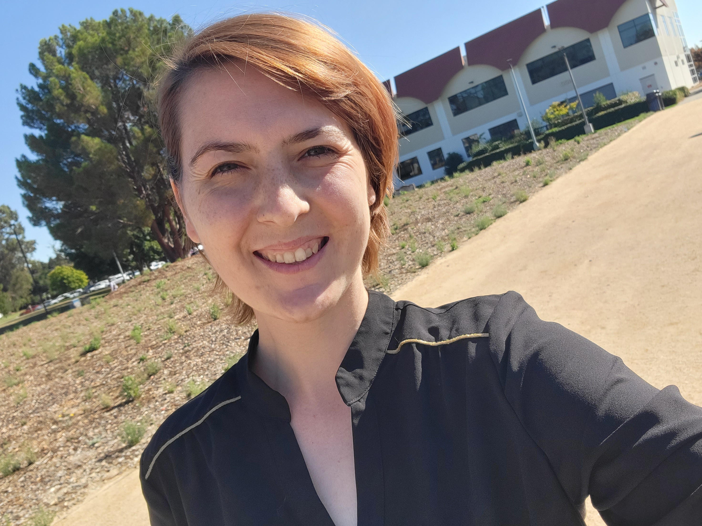 Deshone smiles at the camera in an outdoor selfie, wearing a black collared shirt, with trees and a college campus building in the background on a sunny day.